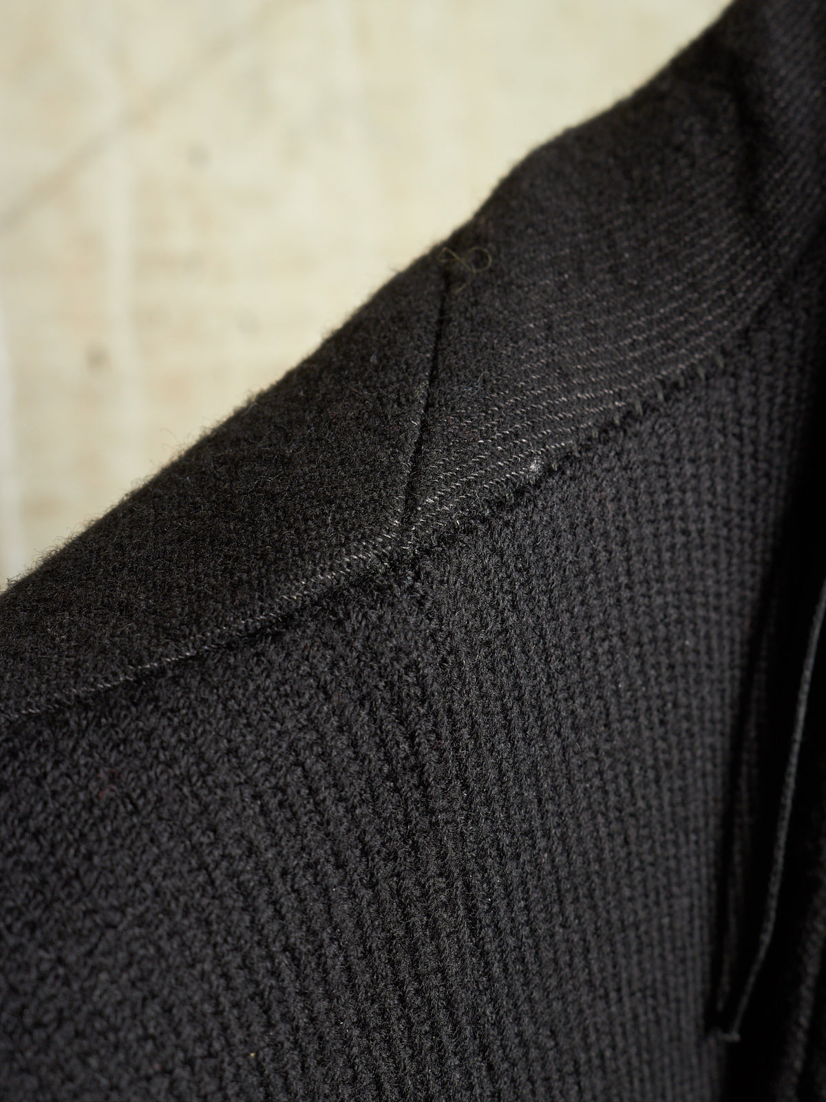 Close-up detail of a black ribbed knit garment, showing the texture and shoulder seam against a light beige background.
