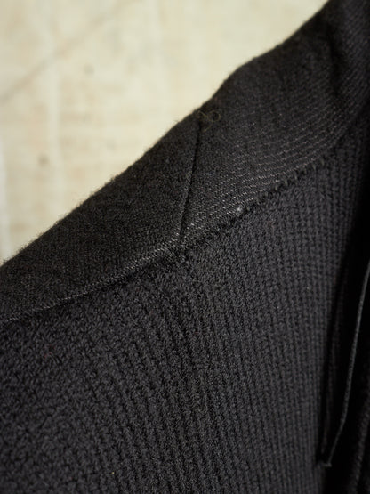 Close-up detail of a black ribbed knit garment, showing the texture and shoulder seam against a light beige background.