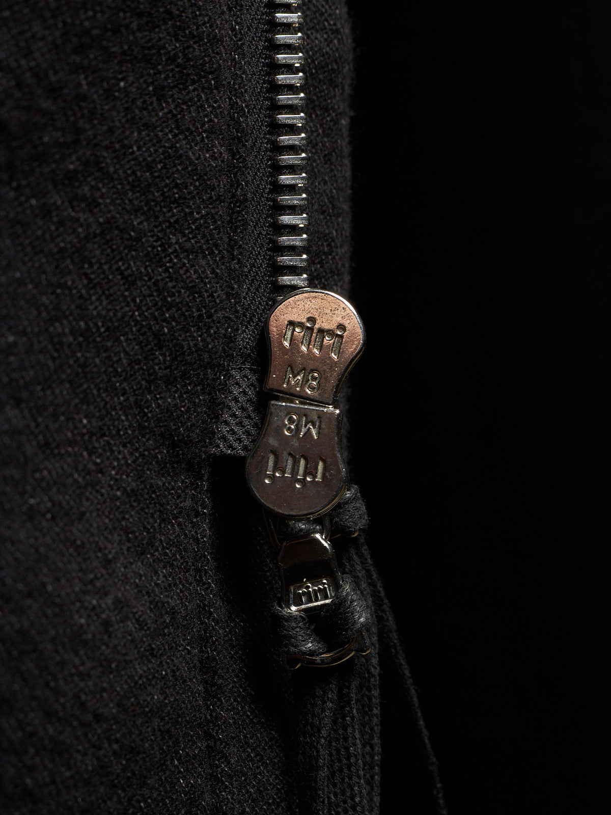 Close-up of a black zippered garment with a bronze-toned metal zipper pull engraved with the brand name 'riti'.