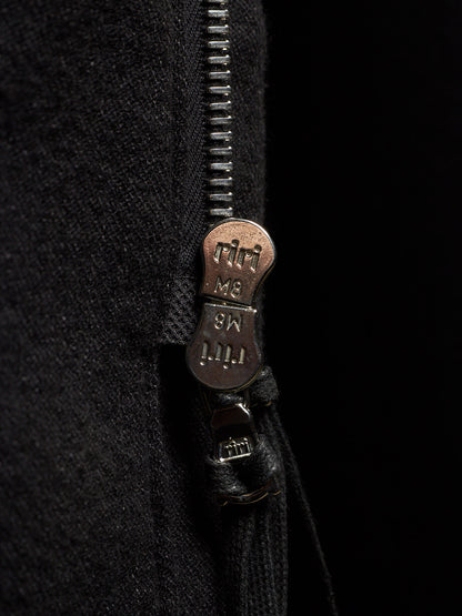 Close-up of a black zippered garment with a bronze-toned metal zipper pull engraved with the brand name 'riti'.