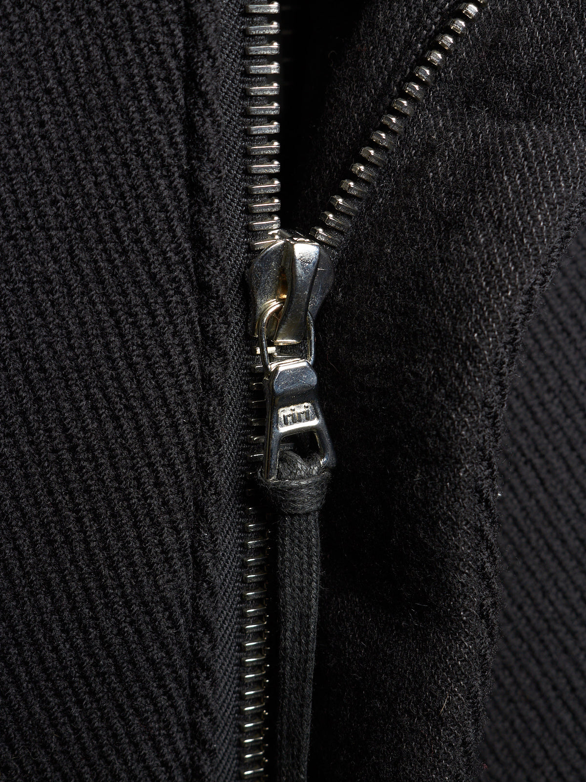 Close-up of a black textured zippered garment with a silver metal zipper pull and black cord lock.