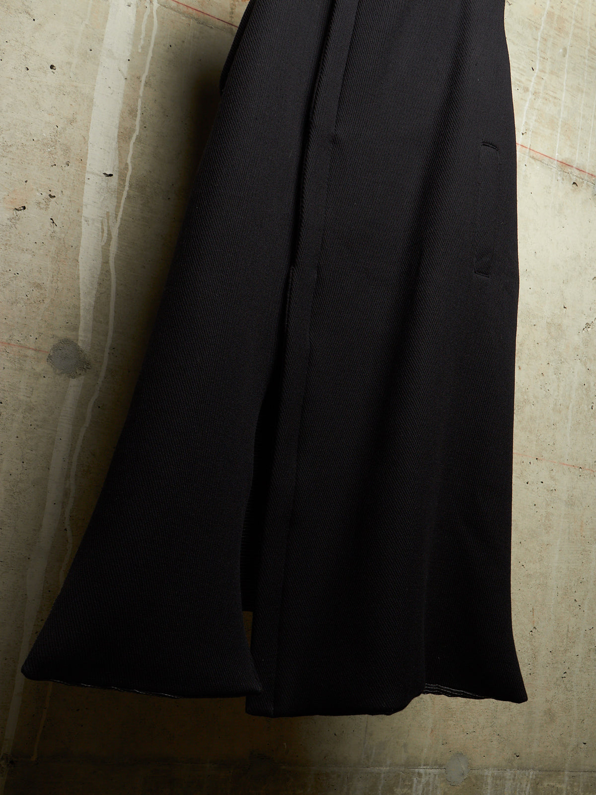 A close-up of a black wide-leg skirt or drape of fabric hanging against a textured concrete wall.