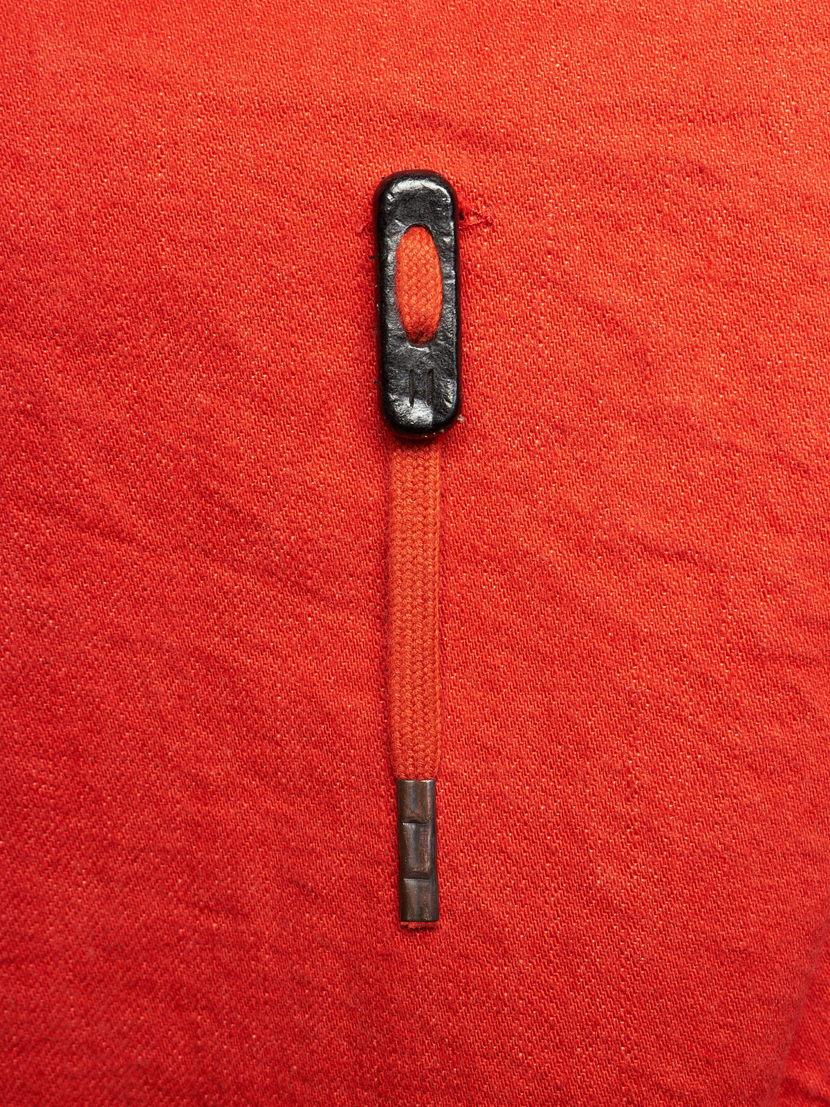 A close-up of a zipper pull with a black metal head and a red-orange zipper tape, shown on a textured red fabric background.