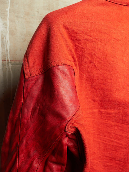Close-up of a red-orange jacket with a textured fabric body and contrasting smooth red leather sleeves, set against a neutral concrete wall.