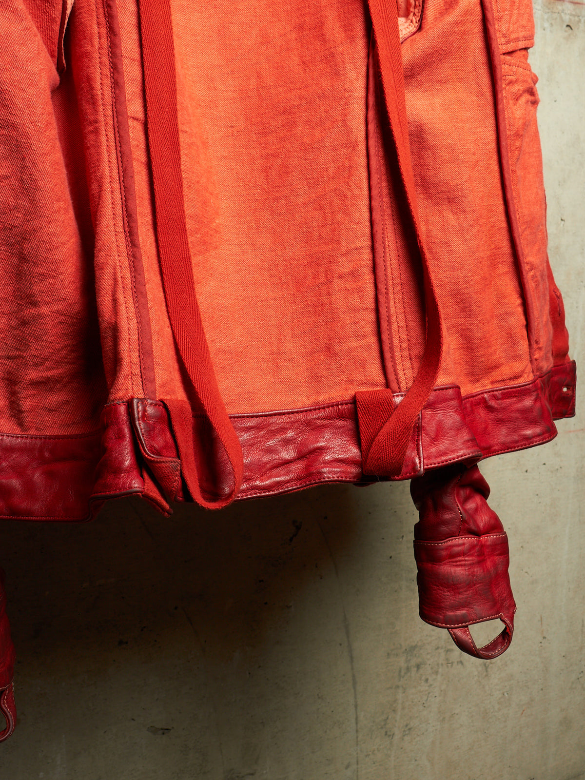 A close-up of a red-orange textured fabric garment or bag with matching red leather trim and straps, hanging against a plain concrete wall.