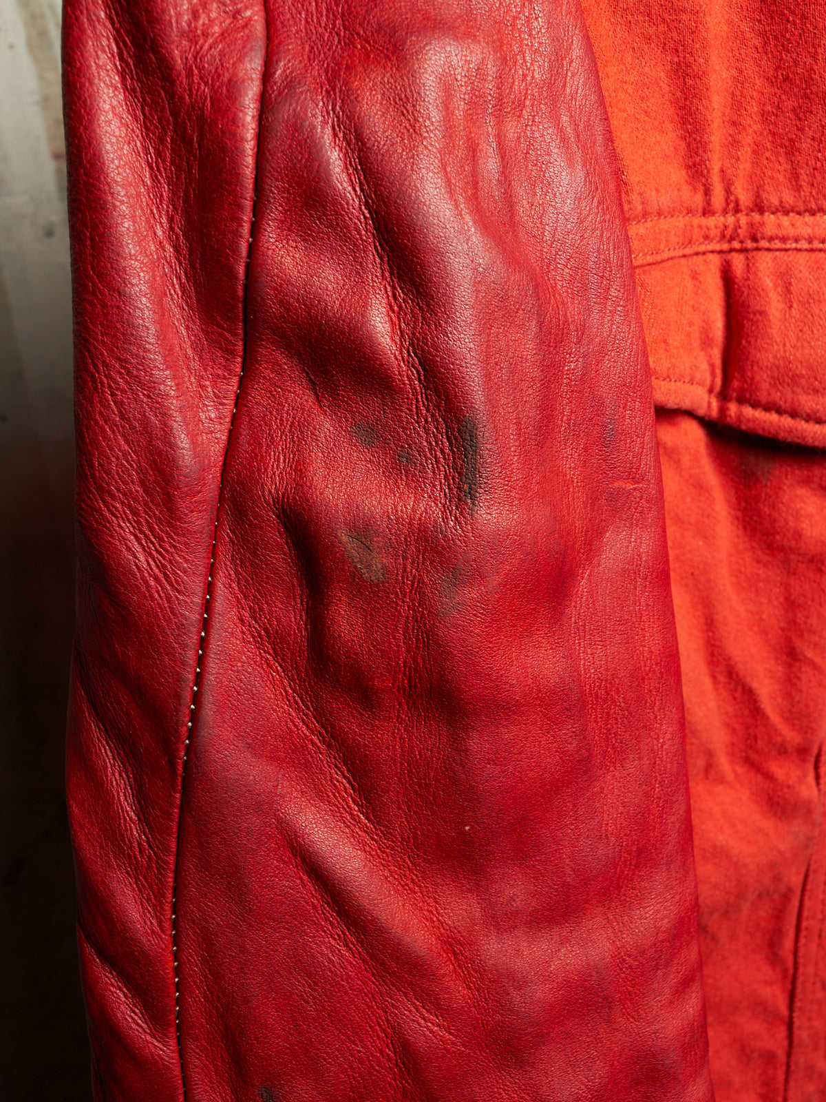 A close-up of a red leather jacket sleeve showing the texture of the leather and some dark stains, next to a bright orange-red fabric section.