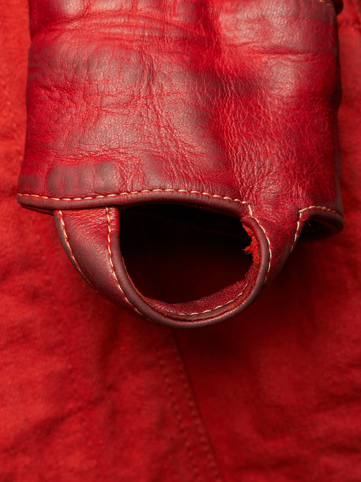 A close-up of a red leather accessory with a textured surface, visible stitching, and a circular cutout or loop detail.