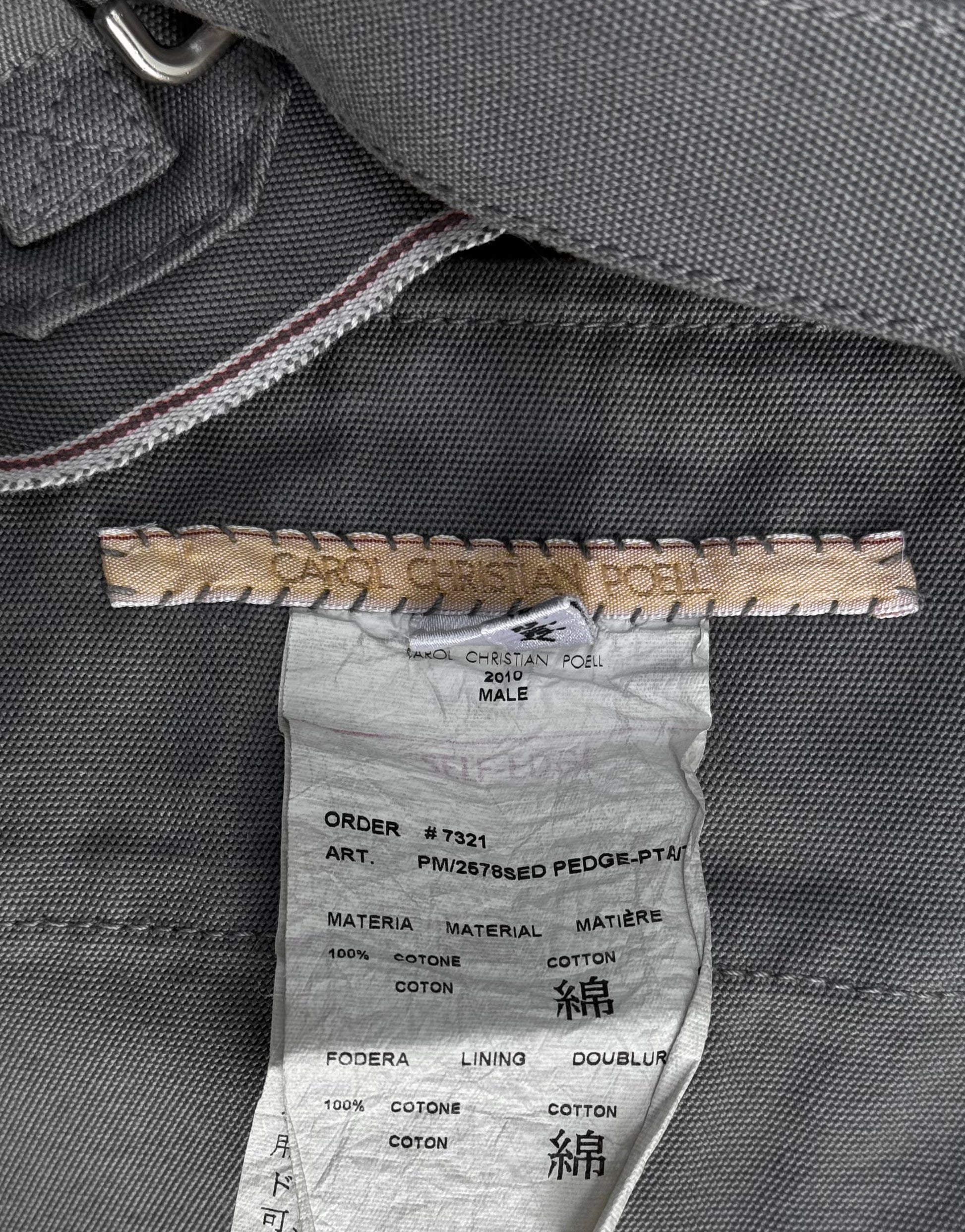 A close-up of a clothing label for Carol Christian Poell’s 2010 “IN-BETWEEN” PM/2578 PEDGE self-edge high-waisted metre pants, showing brand and product details on grey fabric—evoking avant-garde fashion sensibilities.