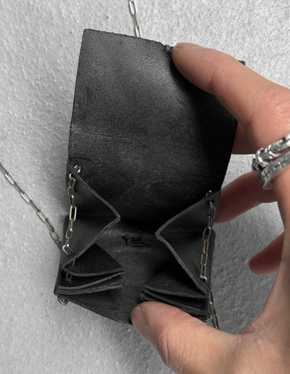 A hand with silver rings holds open the M.A+ by Maurizio Amadei ‘A-B7201’ Calfskin Leather Mini Accordion Pouch Necklace—black leather with chain and stitched pockets—against a textured light gray background. One size fits all essentials.