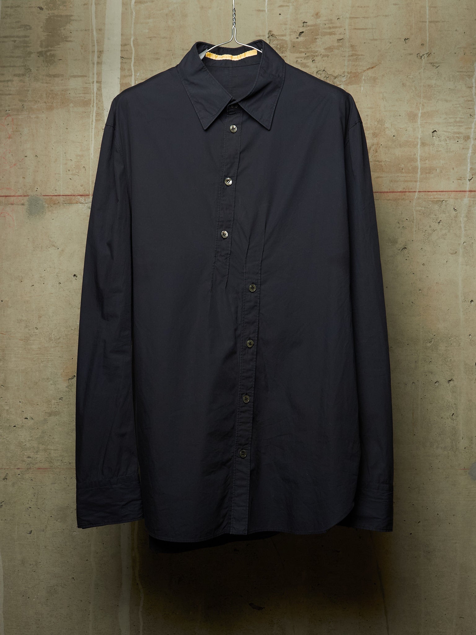 A dark navy blue long-sleeve button-up shirt hanging on a wooden hanger against a textured concrete wall.