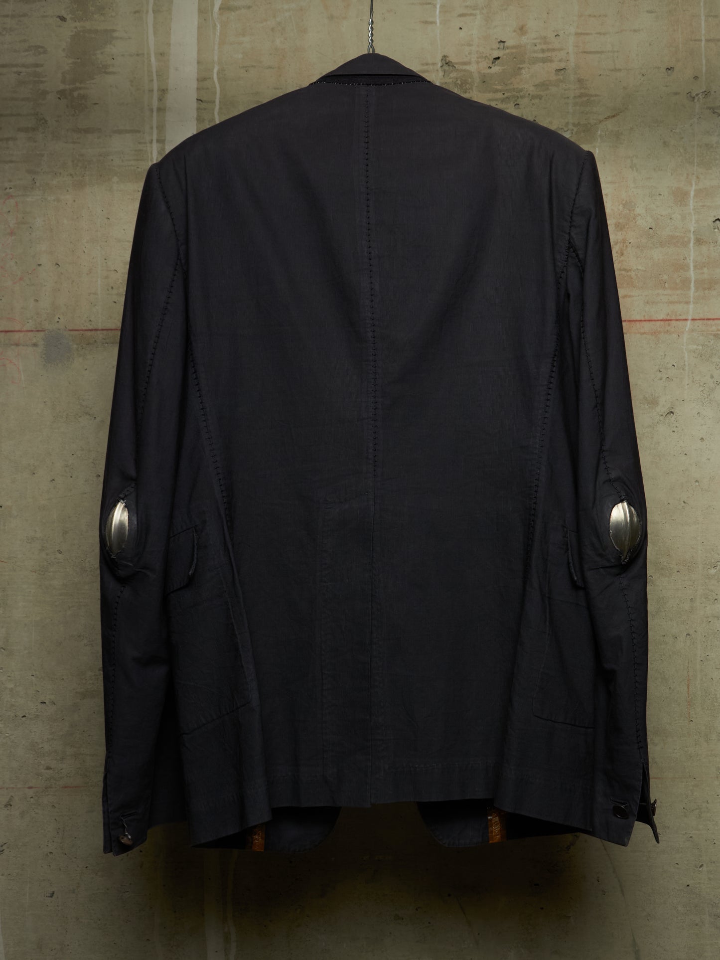 A black jacket hanging against a textured concrete wall, showing the back view with silver metallic elbow patches.