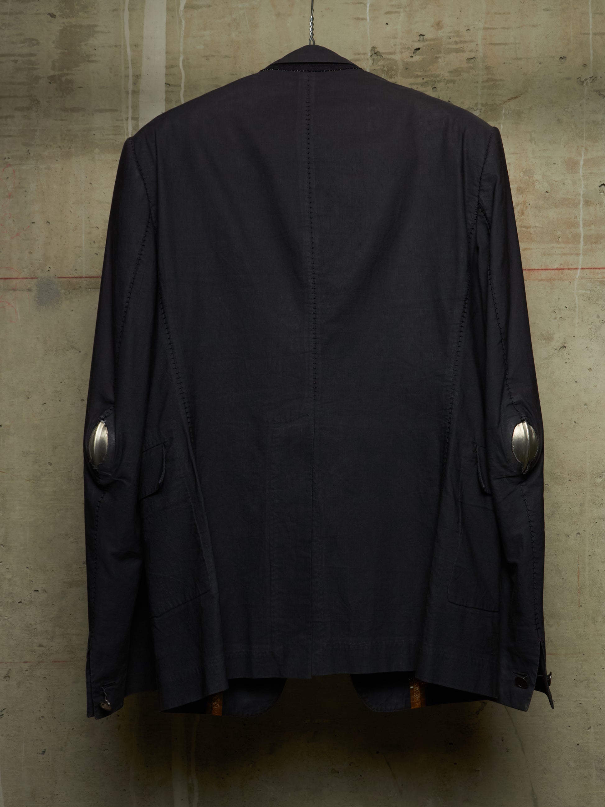 A black jacket hanging against a textured concrete wall, showing the back view with silver metallic elbow patches.