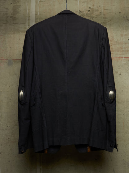 A black jacket hanging against a textured concrete wall, showing the back view with silver metallic elbow patches.
