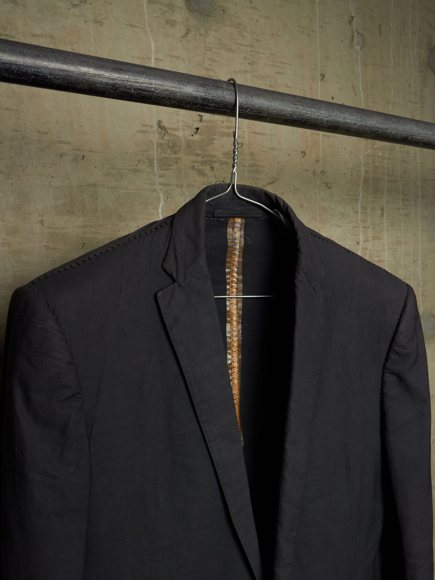 A black tailored blazer jacket hanging on a metal hanger from a dark industrial rod, set against a textured concrete wall.