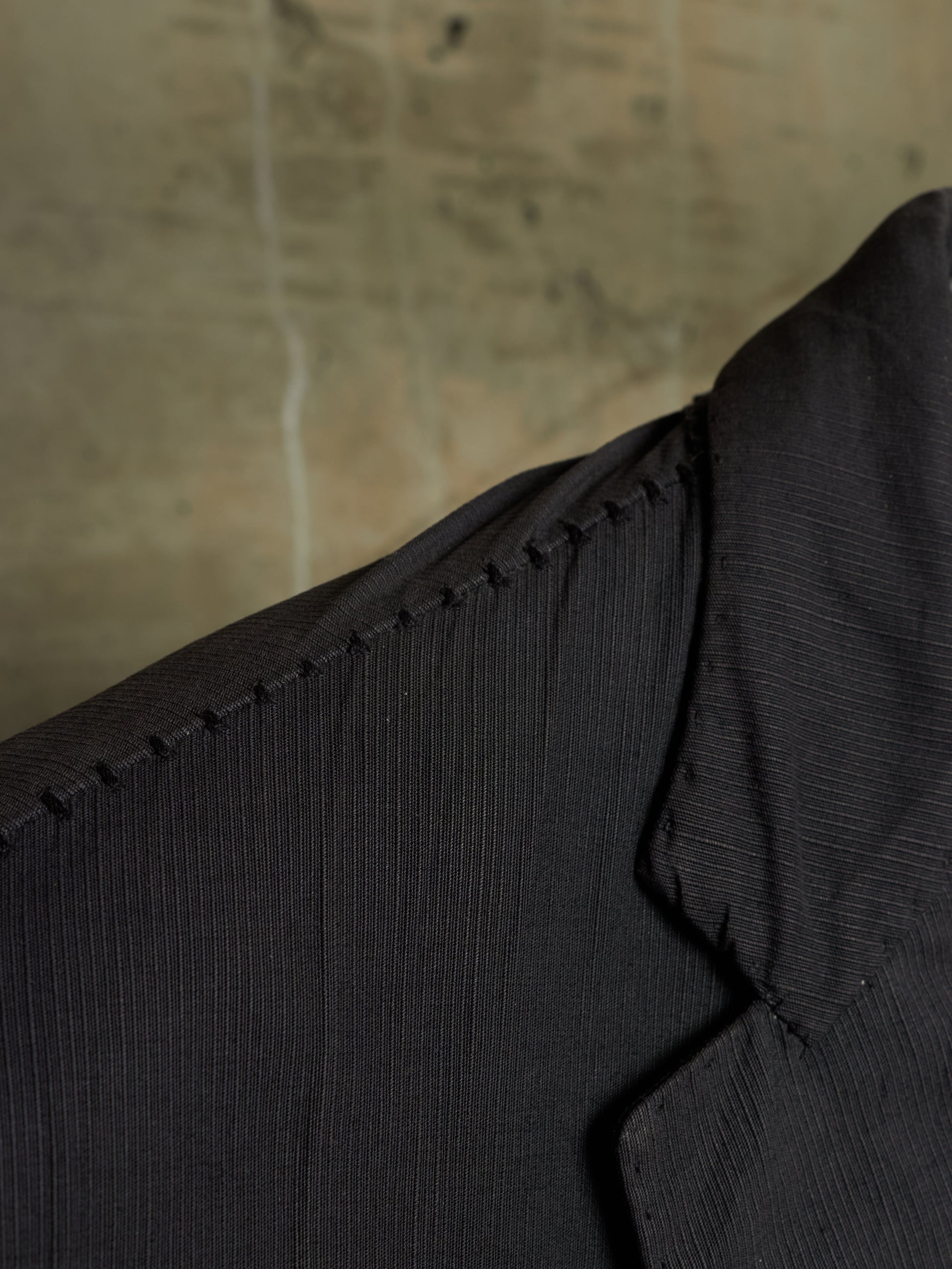 Close-up detail of a black textured jacket or blazer showing the collar, shoulder seam, and fine ribbed fabric against a neutral concrete background.