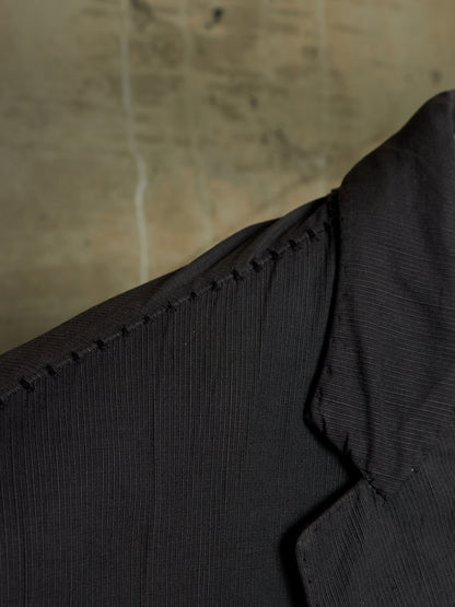 Close-up detail of a black textured jacket or blazer showing the collar, shoulder seam, and fine ribbed fabric against a neutral concrete background.