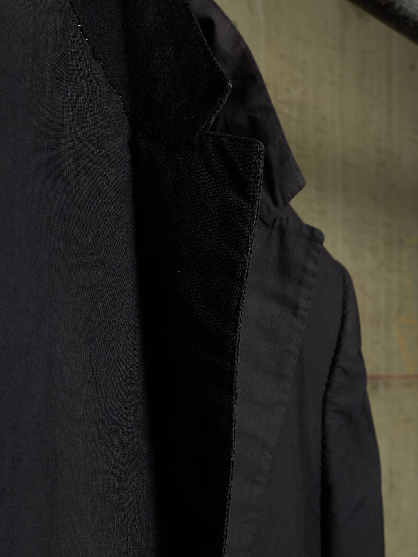 Close-up detail shot of a black jacket, showing the texture of the fabric and the inner lining, set against a neutral textured background.