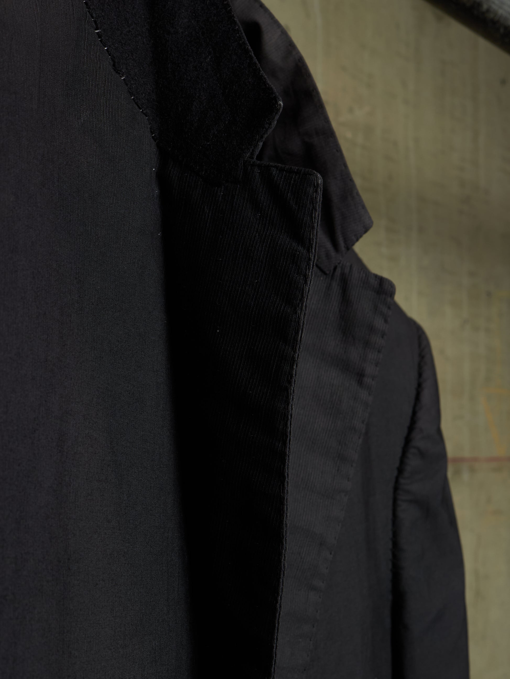 Close-up detail shot of a black jacket, showing the texture of the fabric and the inner lining, set against a neutral textured background.
