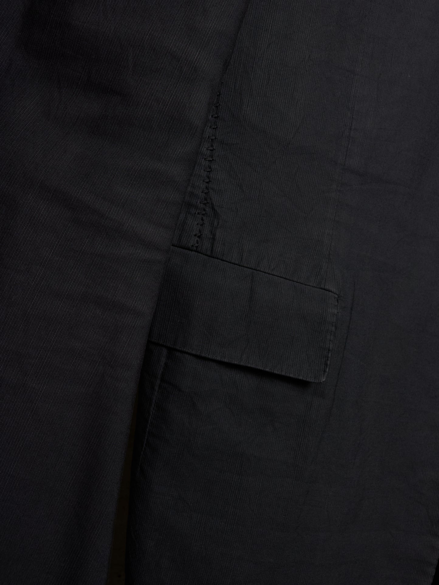 Close-up detail of a black fabric garment showing a side pocket with a flap tab and dark contrast stitching.