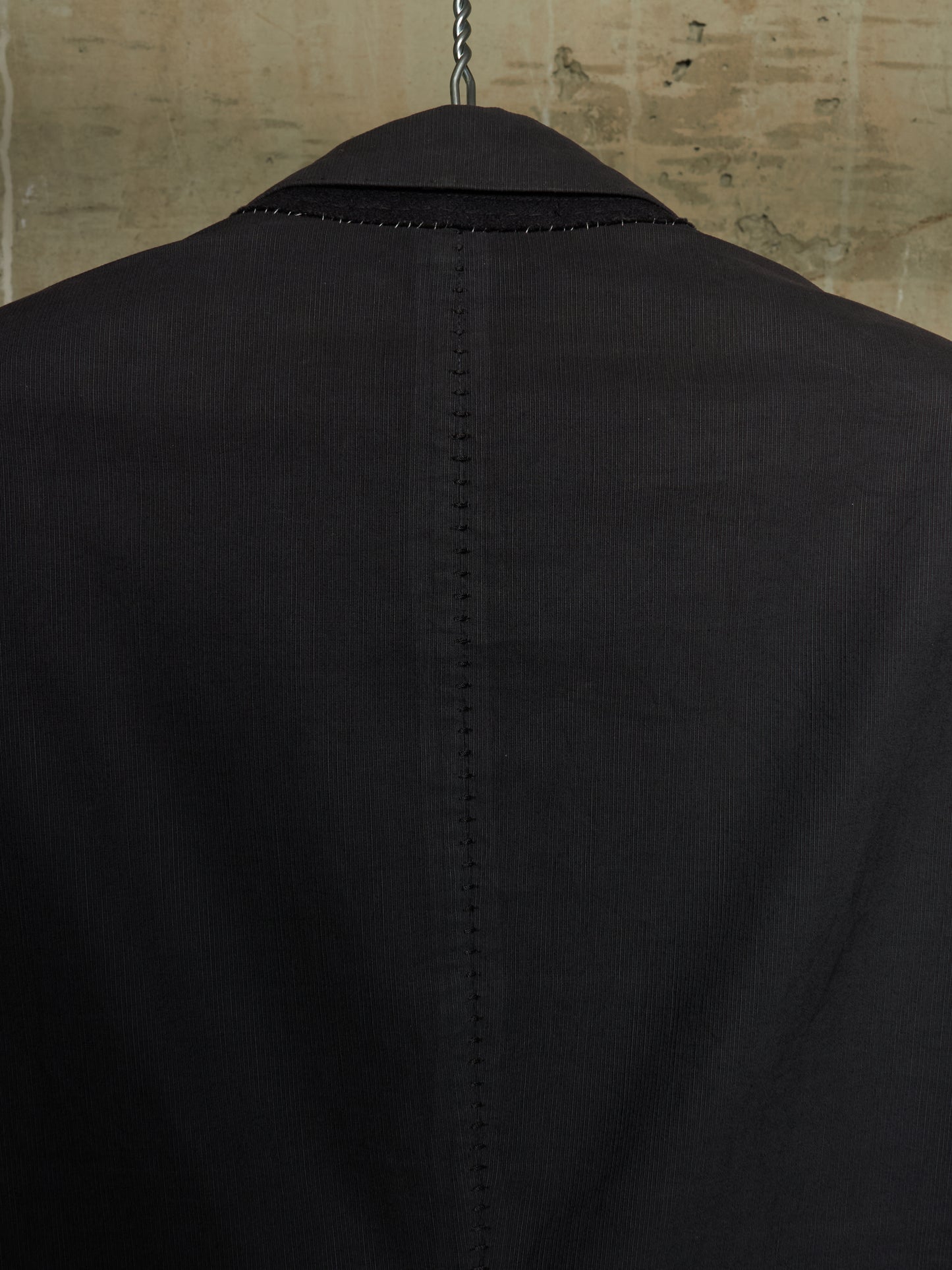 Close-up back view of a black corduroy blazer jacket hanging on a hanger against a textured beige wall.