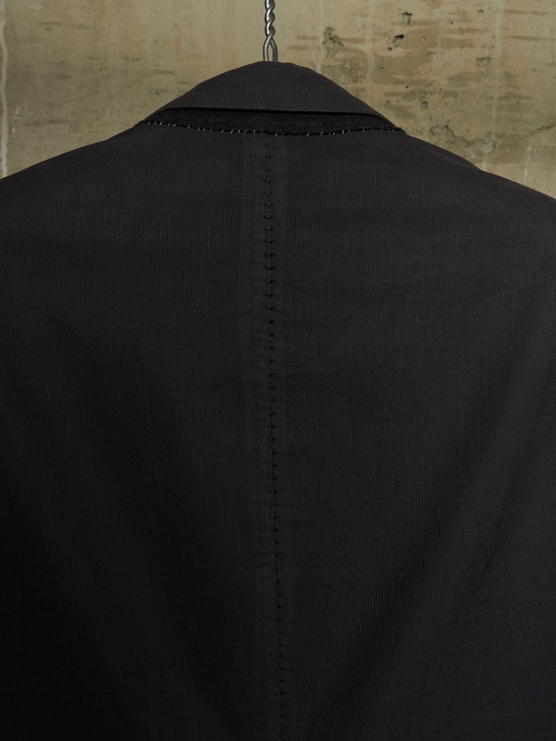 Close-up back view of a black corduroy blazer jacket hanging on a hanger against a textured beige wall.