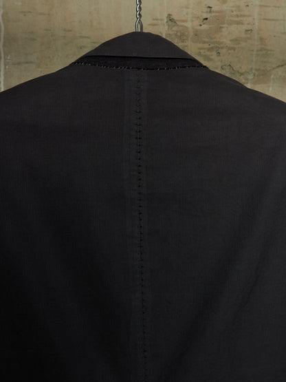 Close-up back view of a black corduroy blazer jacket hanging on a hanger against a textured beige wall.