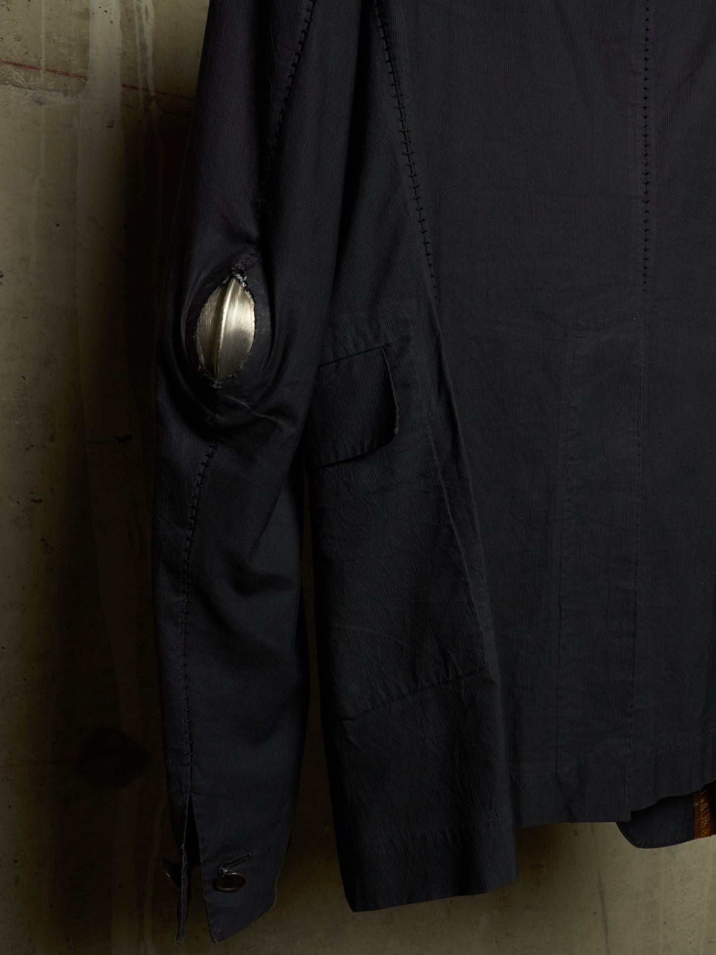 A close-up of a dark navy or black jacket hanging against a textured concrete wall, showing a metallic silver circular patch on the upper sleeve.
