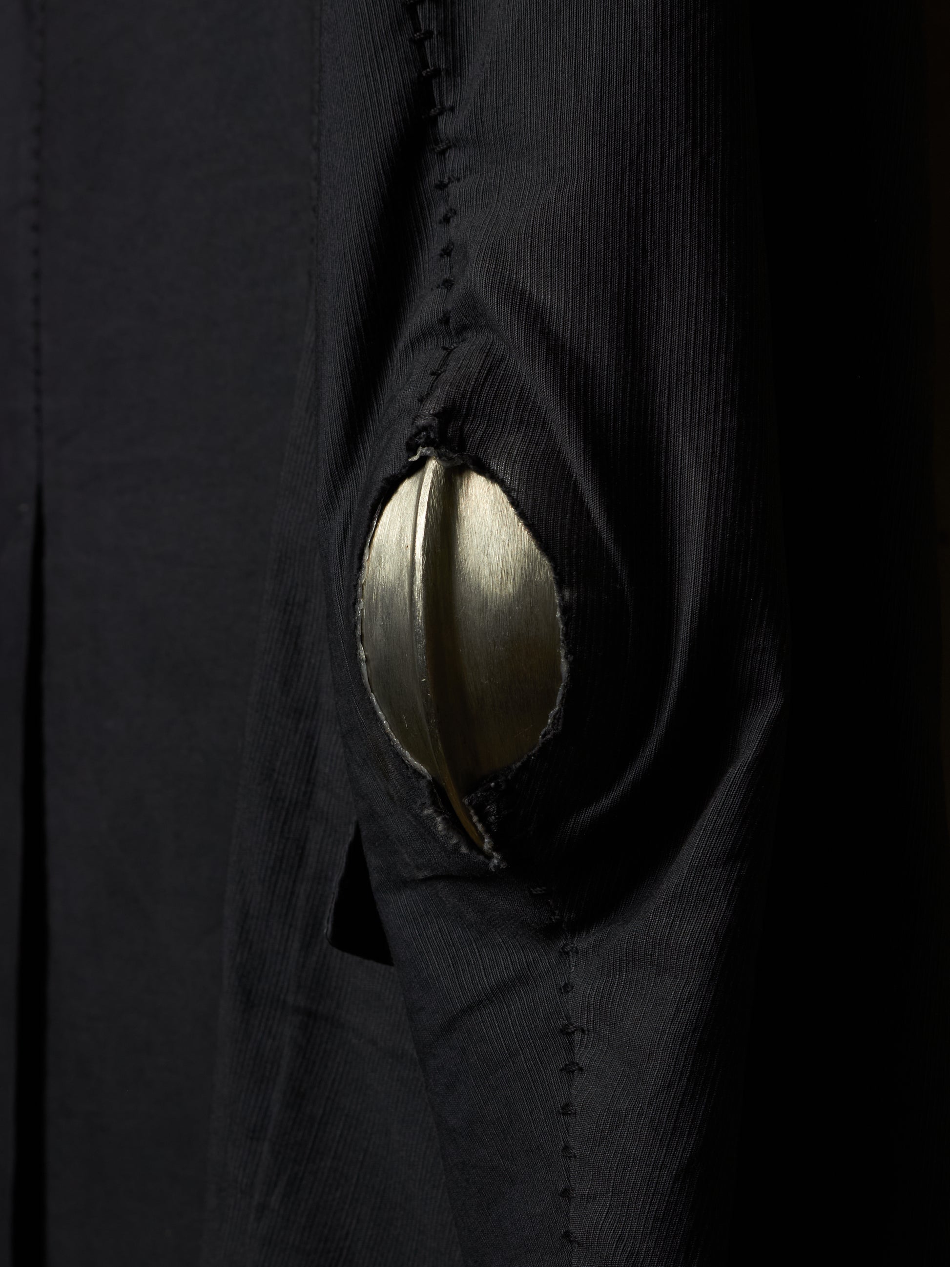 Close-up of a black textured garment with a large metallic gold circular patch or button-like detail on the front.
