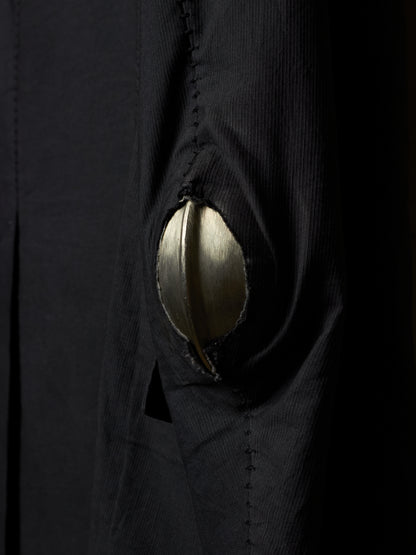 Close-up of a black textured garment with a large metallic gold circular patch or button-like detail on the front.