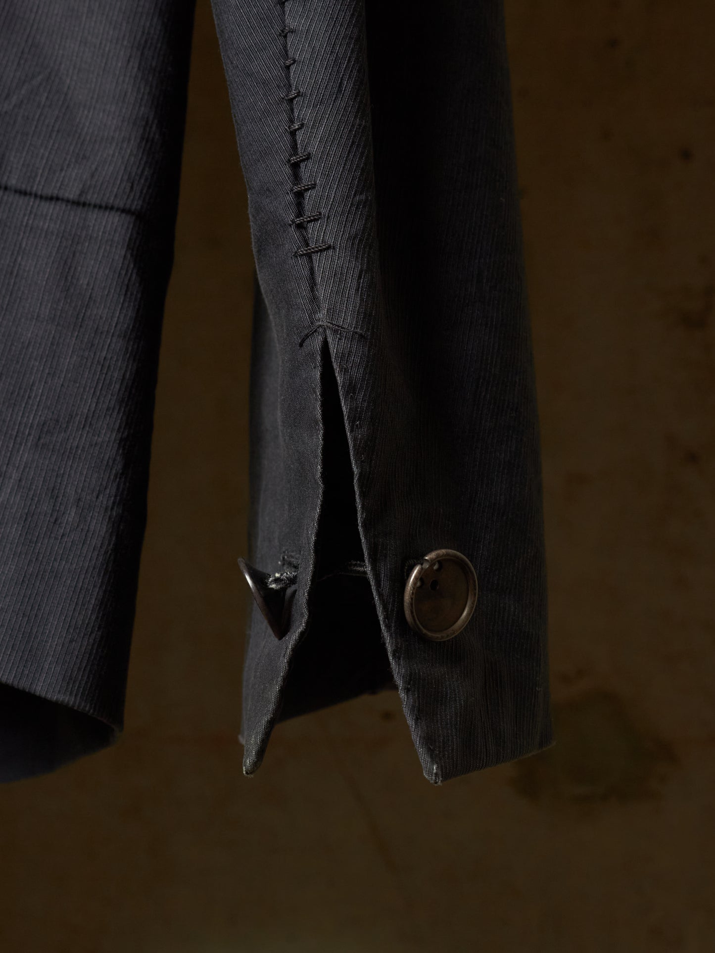 Close-up detail of a dark charcoal gray corduroy jacket sleeve, showing the textured fabric, a decorative button, and a buttonhole with hand-stitched detailing against a warm, dark background.