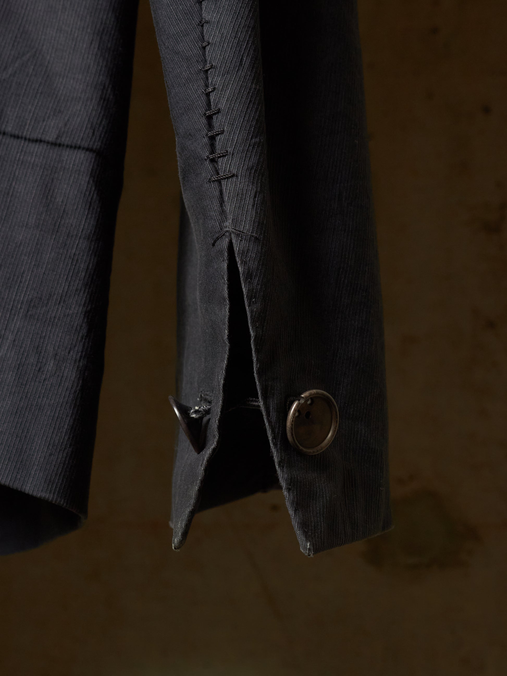Close-up detail of a dark charcoal gray corduroy jacket sleeve, showing the textured fabric, a decorative button, and a buttonhole with hand-stitched detailing against a warm, dark background.