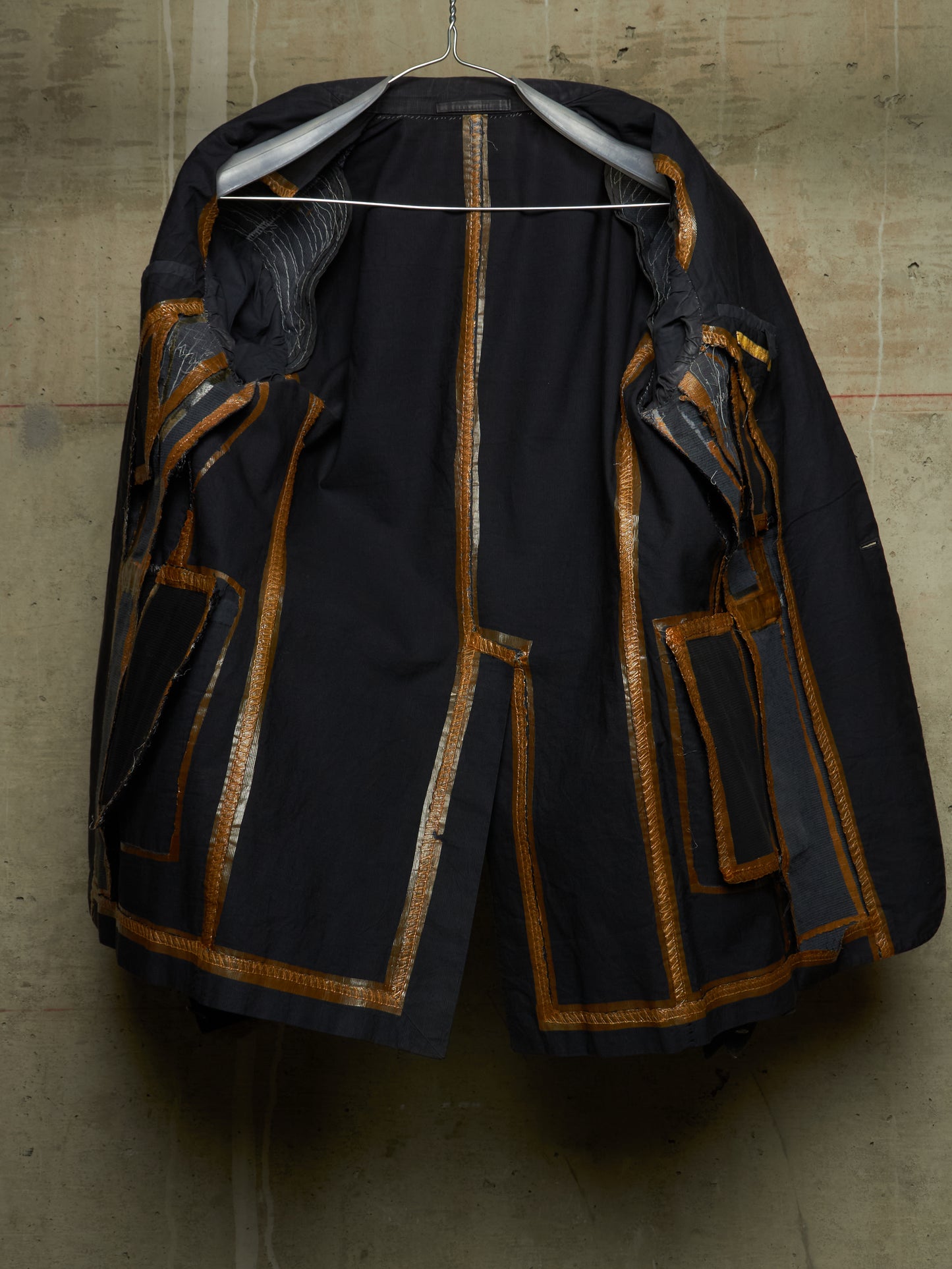 A black vintage jacket with gold metallic trim and patchwork details, hanging on a wire hanger against a textured concrete wall.