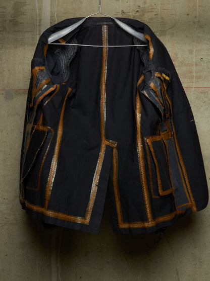 A black vintage jacket with gold metallic trim and patchwork details, hanging on a wire hanger against a textured concrete wall.