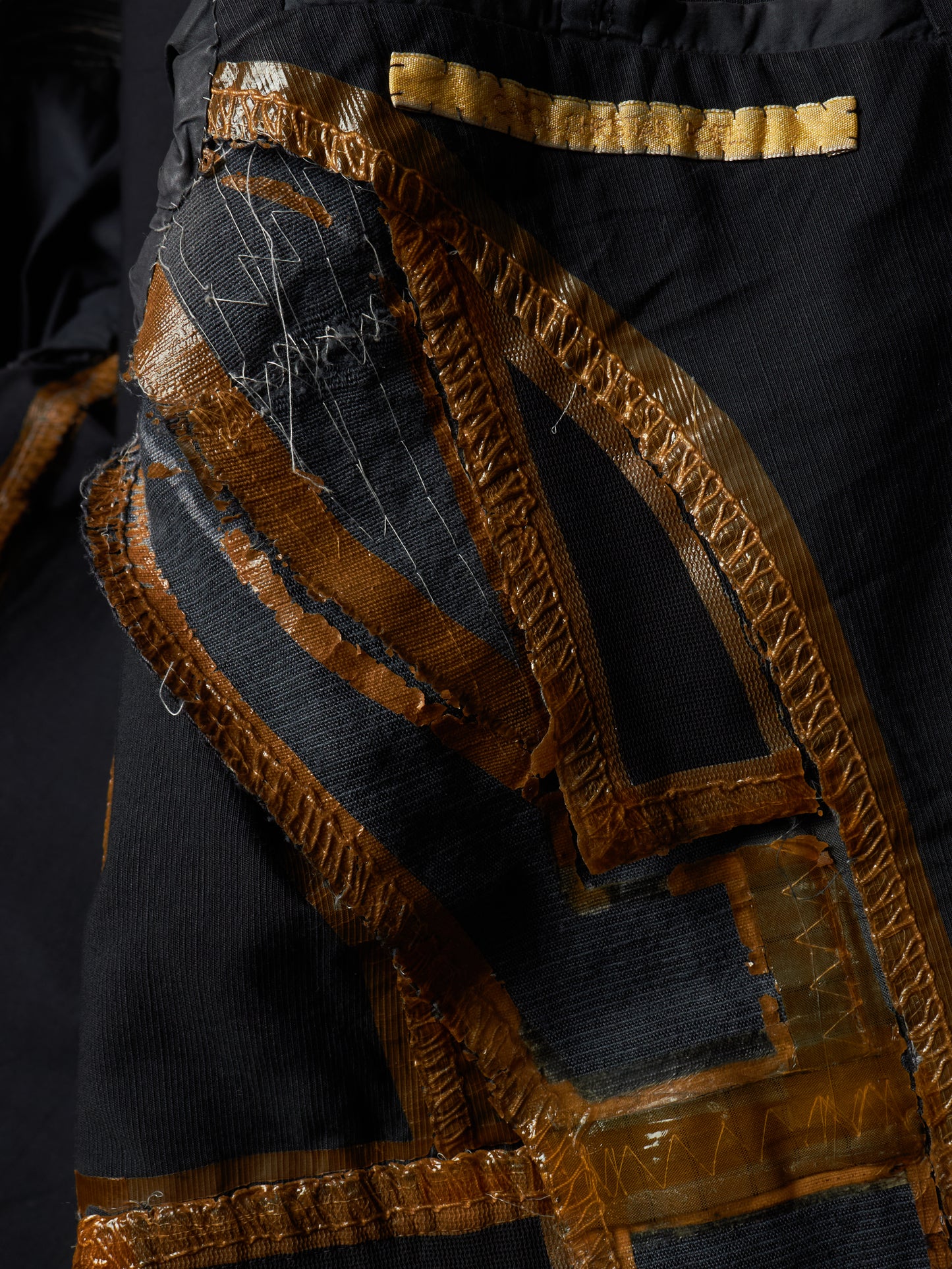Close-up of a black garment featuring large, asymmetrical patches of metallic gold and bronze woven fabric with intricate stitching.