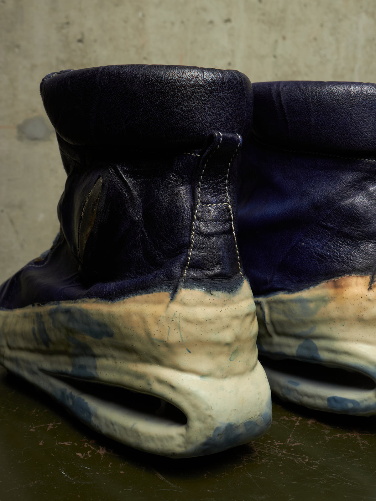 A close-up of a pair of high-top boots with dark navy blue leather uppers and thick, sculptural off-white and blue sculpted soles, photographed against a textured concrete wall.