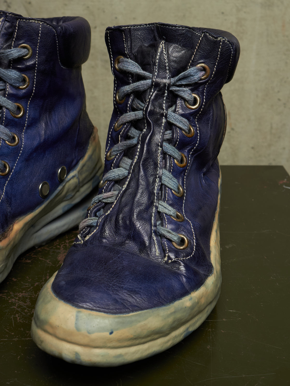 A pair of distressed dark blue leather lace-up boots with heavily worn, scuffed soles and gray laces, displayed on a dark surface against a textured concrete wall.