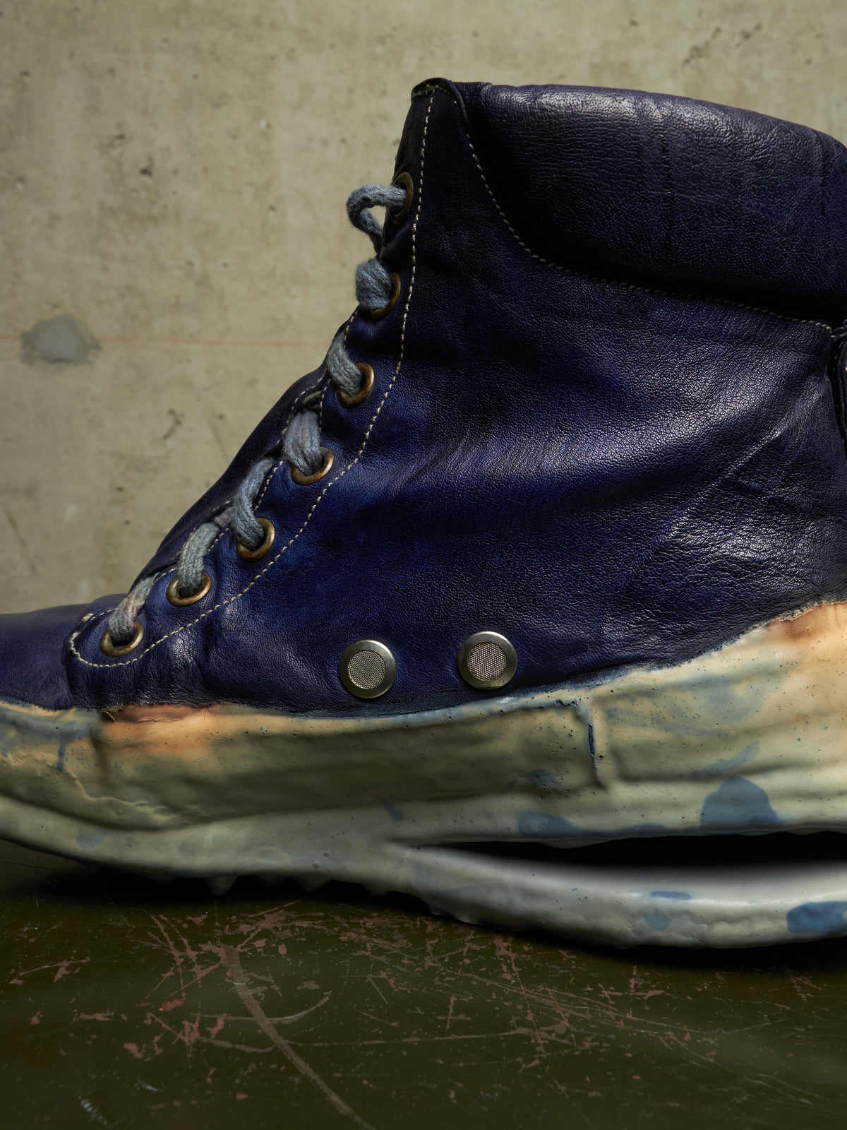 A high-top sneaker boot in dark blue leather with distressed, layered off-white and blue soles, featuring metal eyelets and laces, set against a textured concrete background.