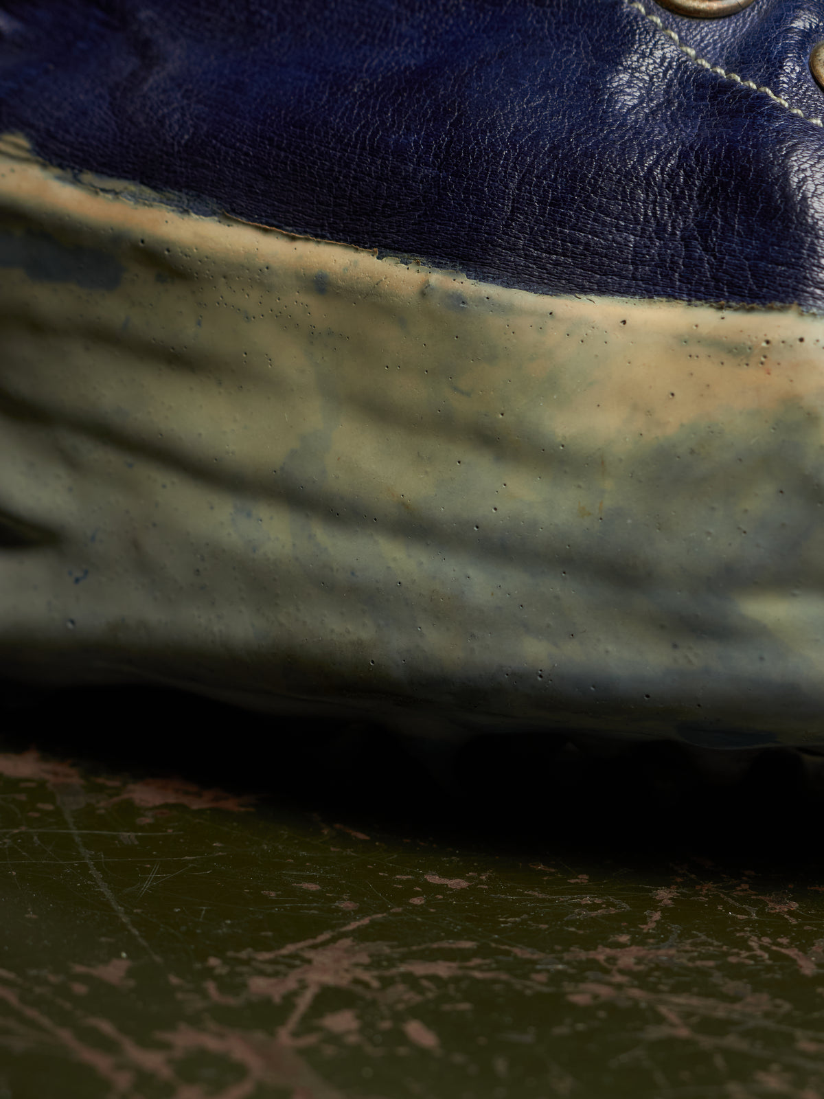 Close-up of a worn blue leather shoe with a heavily scuffed, dirty off-white rubber sole resting on a scratched dark green surface.