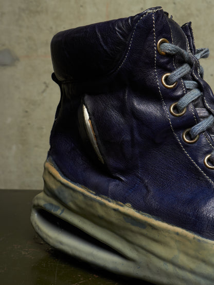 A close-up of a dark navy blue leather high-top shoe with distressed detailing, blue-gray laces, and a thick sculpted off-white sole, shown against a textured concrete background.