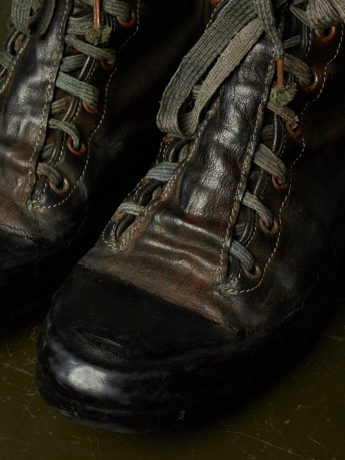 A close-up of a pair of worn black leather lace-up boots with gray laces and brown eyelets, resting on a dark olive-green surface.