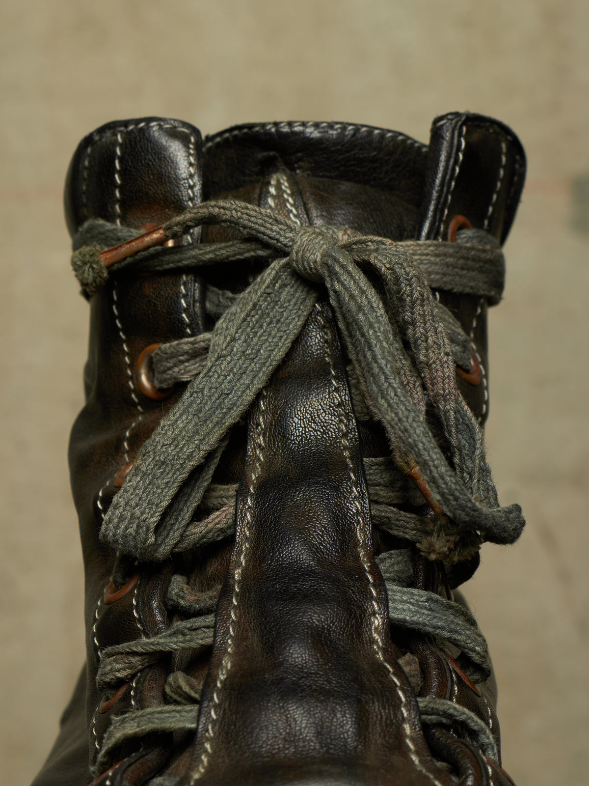 A close-up of a dark brown leather lace-up boot with thick, textured gray laces knotted at the top, set against a neutral beige background.