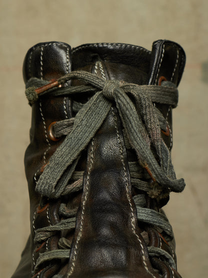 A close-up of a dark brown leather lace-up boot with thick, textured gray laces knotted at the top, set against a neutral beige background.