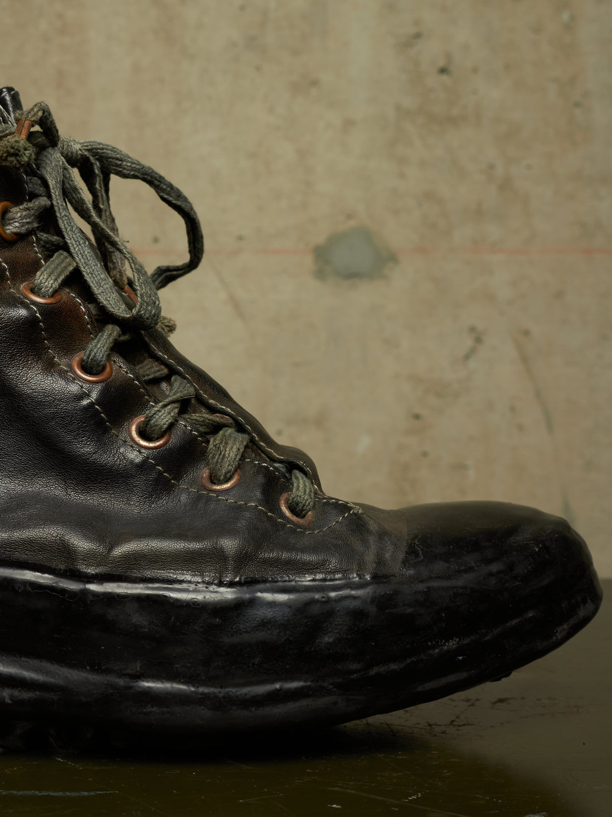 A close-up of a worn black leather lace-up boot with dark laces and copper-toned eyelets, set against a textured beige concrete wall.
