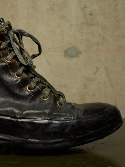 A close-up of a worn black leather lace-up boot with dark laces and copper-toned eyelets, set against a textured beige concrete wall.