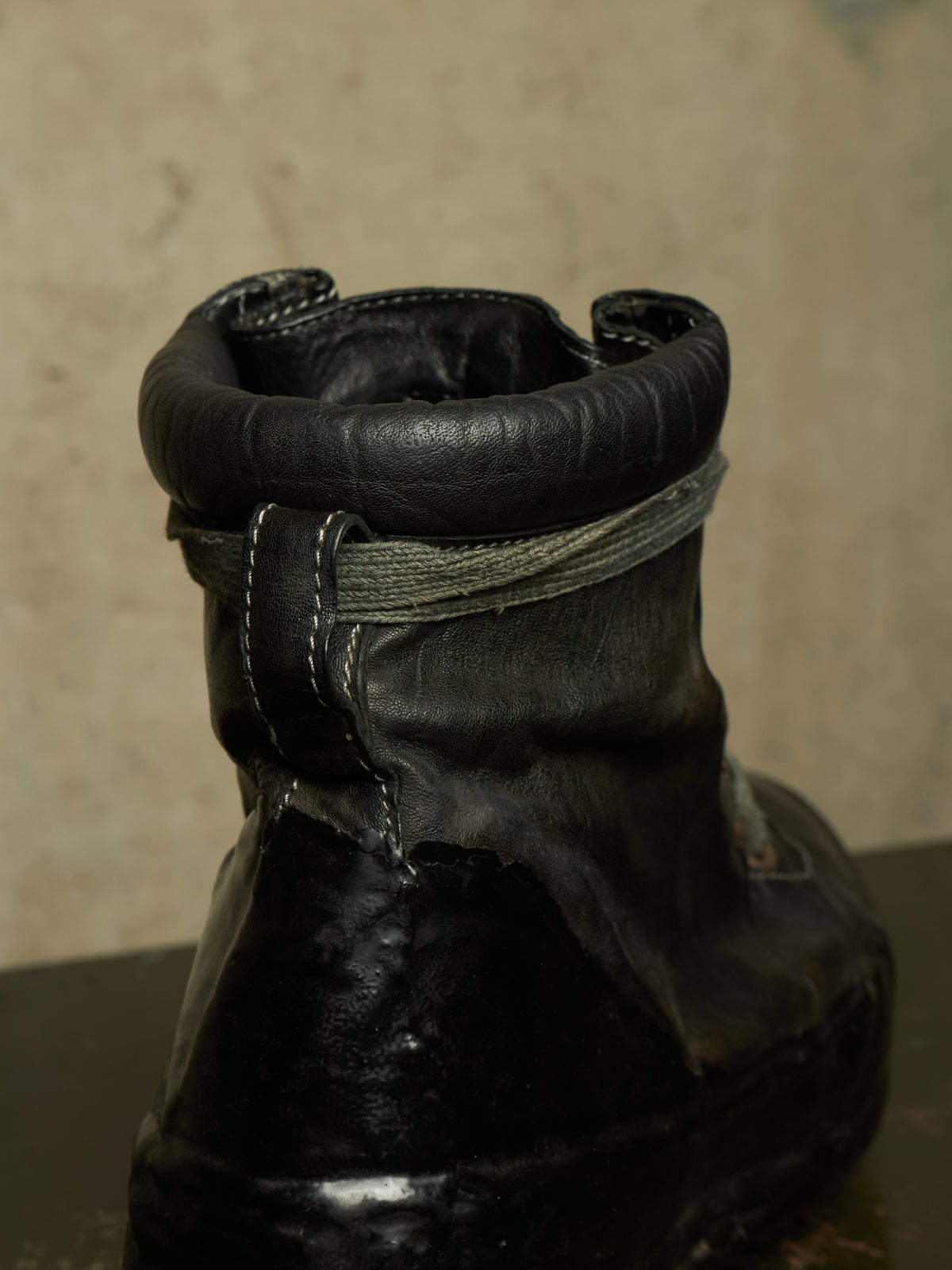 A close-up of a worn black leather boot with a thick black sole, showing a frayed, distressed texture and a gray fabric strap around the collar, set against a textured beige background.
