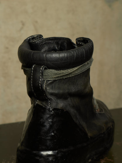 A close-up of a worn black leather boot with a thick black sole, showing a frayed, distressed texture and a gray fabric strap around the collar, set against a textured beige background.