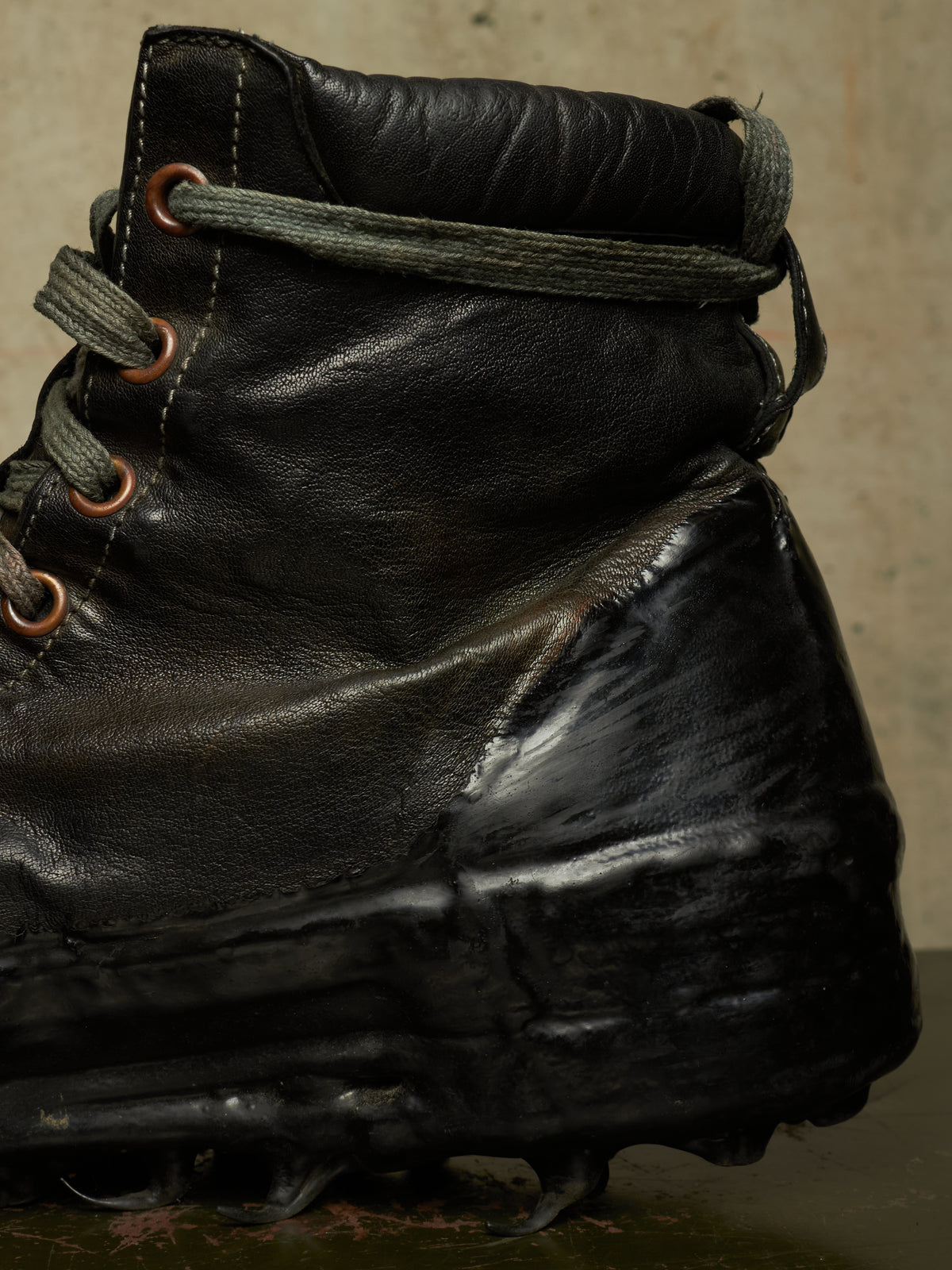 A close-up of a black leather high-top boot with thick gray laces and metal eyelets, photographed against a textured beige background.