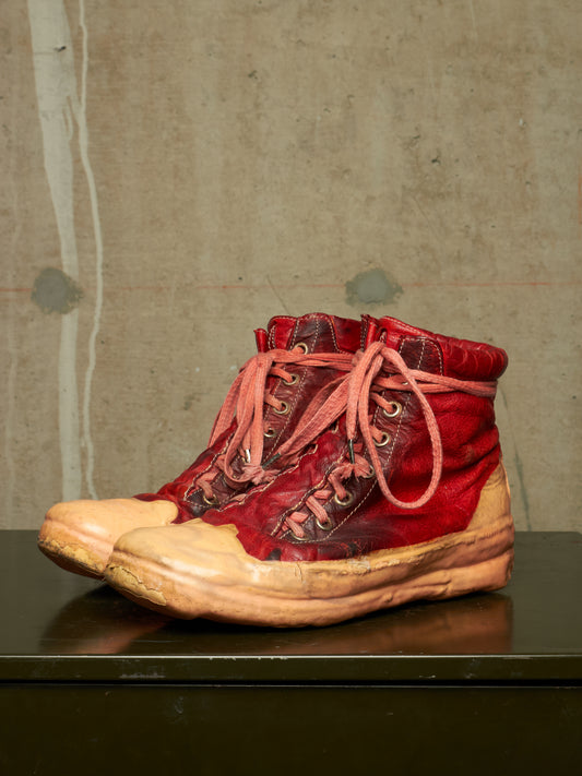 A pair of high-top red leather boots with tan rubber toe caps and matching red laces, displayed on a dark surface against a textured concrete wall.