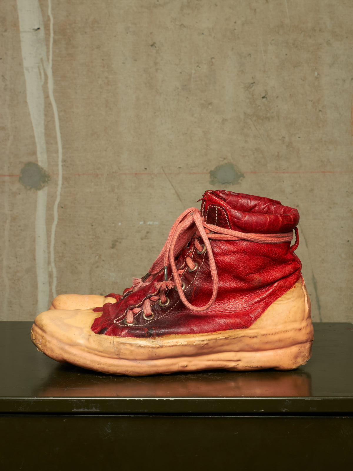A single high-top sneaker with a distressed red leather upper and a tan rubber sole, resting on a dark reflective surface against a textured concrete wall.