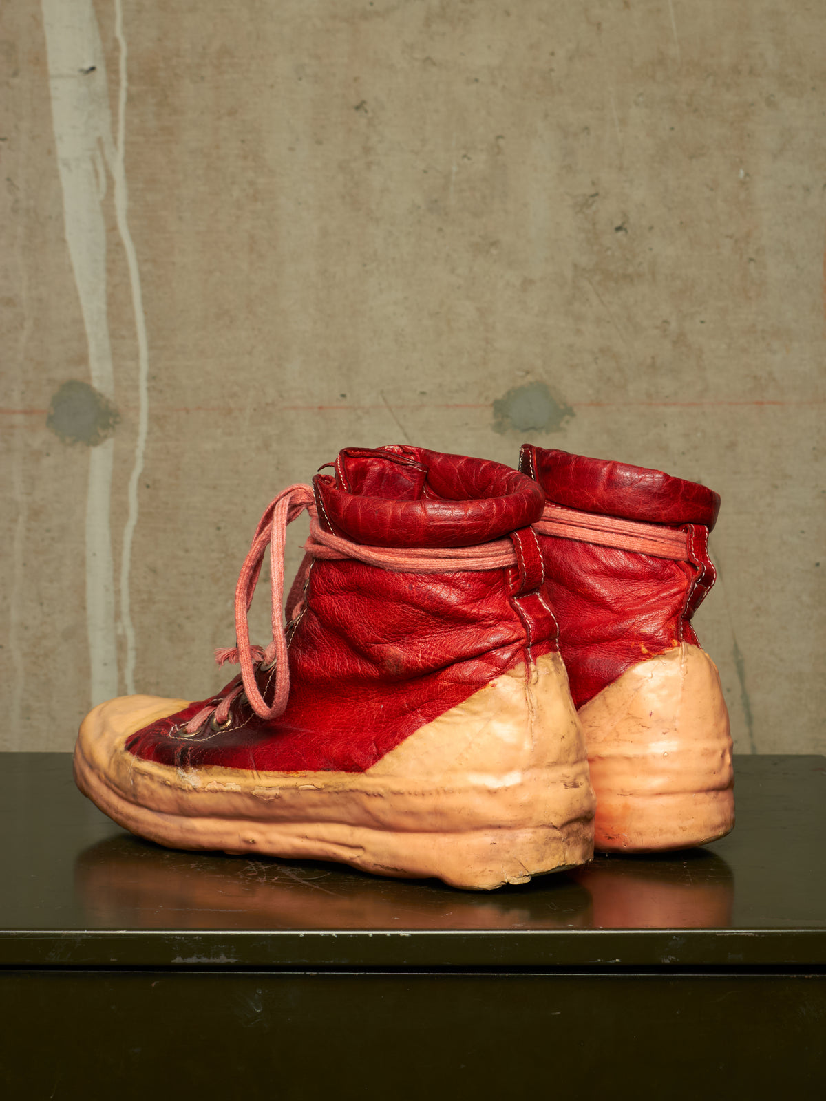 A pair of high-top sneakers with bright red leather uppers and thick beige rubber soles, displayed on a dark reflective surface against a textured concrete wall.